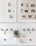 Double bathroom sink with chrome faucets, white subway tiles, small potted plant, and framed photos on the wall.