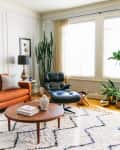 Mid-century modern living room with orange sofa, black lounge chair, large plants, and a round wooden coffee table.