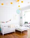 Nursery with white crib, toddler bed, colorful pom-pom garland, and yellow and white hanging decorations.