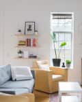Bright living room with gray sofa, yellow armchair, wall shelves with books, plants, and framed art.