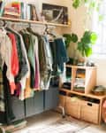 Clothing rack with colorful clothes, books on a shelf, potted plant, and woven baskets in a bright room.