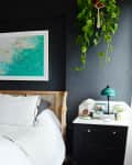 Bedroom with dark walls, white bedding, a rattan headboard, hanging plant, and a turquoise beach painting above the bed.