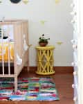 Nursery with wooden crib, colorful patchwork rug, yellow side table, and potted plant against a wall with bee decals.
