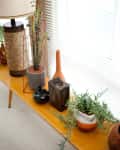 Wooden console table with potted plants, ceramic vases, and a lamp near window blinds.