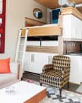 Loft bed with ladder, geometric armchair, and modern decor in a compact living space with a kitchenette.