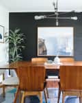 Dining room with wooden table, six chairs, black accent wall, large framed artwork, and a potted plant.