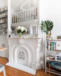 White kitchen with wooden countertop, ornate fireplace, open shelves, and a small table with books and a plant.