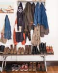Coat rack with various jackets and coats above a bench lined with boots and shoes, wicker baskets on top shelf.