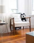 Wooden armchair with white cushions, striped pillow, and a black-and-white plush cat toy in a bright room.