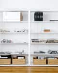 White shelving unit with wire baskets, books, and storage boxes, next to a potted plant and a wastebasket with rolled papers.