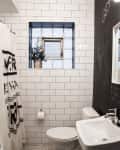 Small bathroom with white subway tiles, a window with a potted plant, toilet, sink, and a blackboard wall with writing.