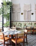 Restaurant interior with macramé wall hangings, patterned floor tiles, wooden chairs, and floral cushioned seating.