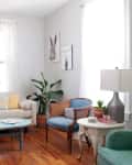 Bright living room with a beige sofa, blue armchair, plants, and colorful artwork on the walls.