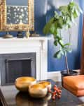 Gold bowls and orange decor on a dark table, with a fiddle leaf fig, ornate mirror, and blue walls in the background.