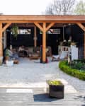 Covered patio with wooden beams, outdoor seating, potted plants, and gravel flooring in a garden setting.