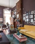 Living room with yellow sofa, gray armchair, pink ottoman, blue rug, and gallery wall on exposed brick.