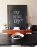 Chalkboard with "Eat Drink Enjoy" above a wooden sideboard, vase of flowers, and a bowl on a dark dining table.