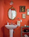 Small bathroom with orange patterned wallpaper, pedestal sink, gold-framed mirror, and teal shelving with towels and decor.