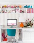 Home office with white desk, teal chair, orange lamp, computer, and colorful decor on shelves.