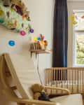 Cozy nursery with a wooden crib, colorful string lights, plush toys, and a chair by a large window with dark curtains.