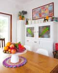 Dining room with wooden table, fruit centerpiece, white cabinet with stained glass, and colorful wall art.