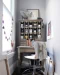 Small home office with a wooden desk, white chair, cubby shelf, and art supplies by a window.