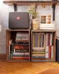 Books and decor on wooden crates, with a vintage speaker, potted plant, and yellow velvet chair in a cozy room.