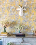 Yellow floral wallpaper with a white deer head, marble mantel, books, vases of flowers, and brass candlesticks.