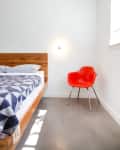 Minimalist bedroom with a wooden bed, geometric blanket, and a red chair under a wall light.