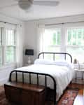 Bedroom with black metal bed, white bedding, wooden chest, potted plants, and large windows with cream curtains.