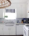 White kitchen with marble countertops, striped window shade, wicker light fixture, and appliances including a mixer and kettle.