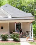 Front porch of a beige house with a mint green door, red trim, rose bushes, and two people sitting with a dog.