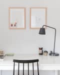 Minimalist workspace with a white desk, black chair, desk lamp, and framed art on the wall.