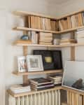 Corner bookshelf with stacked books, framed art, a small speaker, and decorative items on light wood shelves.