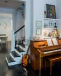 Wooden piano with sheet music, guitar, and music stand beside a staircase in a cozy living room.