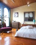 Bedroom with wooden ceiling, large window, red chair, wooden dresser, bed with white linens, and a cat on the floor.