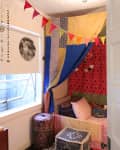 Cozy nook with colorful patchwork canopy, patterned cushions, and bunting flags, next to a window with a decorative blind.