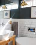 Bathroom with white subway tiles, a wooden vanity, round mirror, brass pendant lights, and abstract wall art.