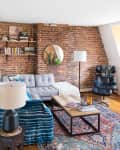 Cozy living room with exposed brick wall, gray sofa, leather chair, round mirror, and colorful patterned rug.