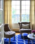 Two tufted chairs with a decorative pillow on a blue patterned rug in a sunlit room with large windows and beige curtains.