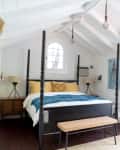 Bright bedroom with a four-poster bed, yellow pillows, blue throw, wooden bench, and large windows with sheer curtains.