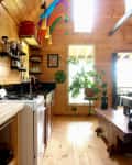 Wooden kitchen with open shelves, colorful hanging decor, potted plant, and a rustic bench under a skylight.