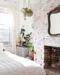 Bright bedroom with white bedding, brick fireplace, hanging plants, and large windows with sheer curtains.