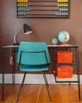 Teal chair at a desk with a black lamp, globes, orange boxes, and a colorful abacus on a brown wall.