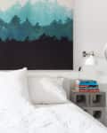 Bed with white linens, forest-themed artwork above, concrete block nightstands, books, and wall-mounted lamps.