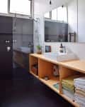Modern bathroom with a glass shower, wooden shelves, white sink, potted plant, and neatly stacked towels.