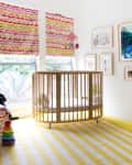 Nursery with a round wooden crib, colorful blinds, striped yellow rug, and wall art.