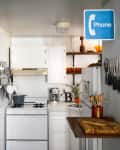 Small kitchen with white cabinets, brick wall, stove, cutting board, and a blue phone sign.