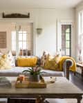 Living room with a gray sofa, yellow and floral pillows, wooden coffee table, and large windows letting in natural light.
