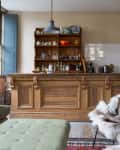 Cozy kitchen with wooden island, open shelves, green sofa, fur-covered chair, and large windows with blue shutters.